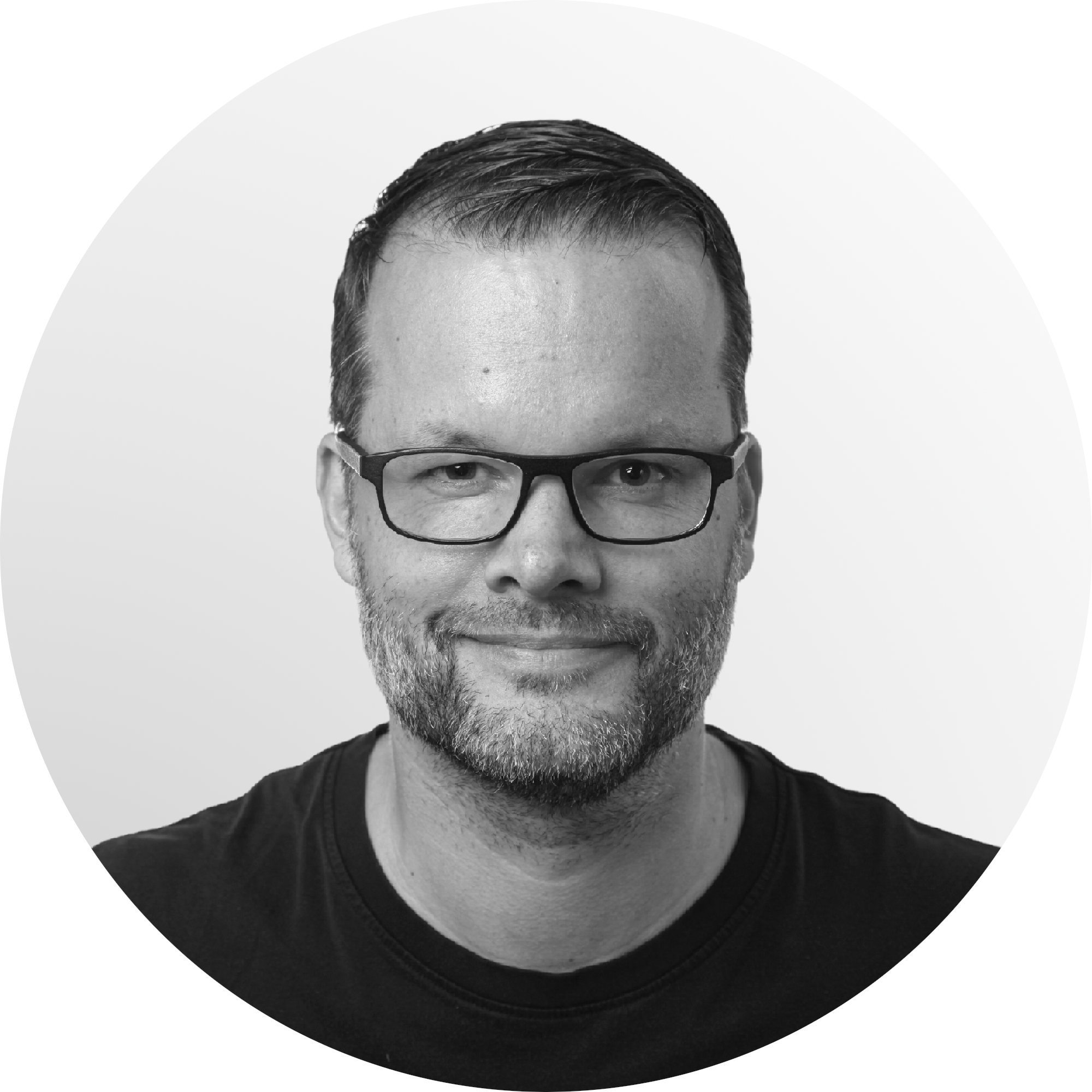 Black and white portrait of a smiling man with glasses and a beard, wearing a black t-shirt, against a plain background.