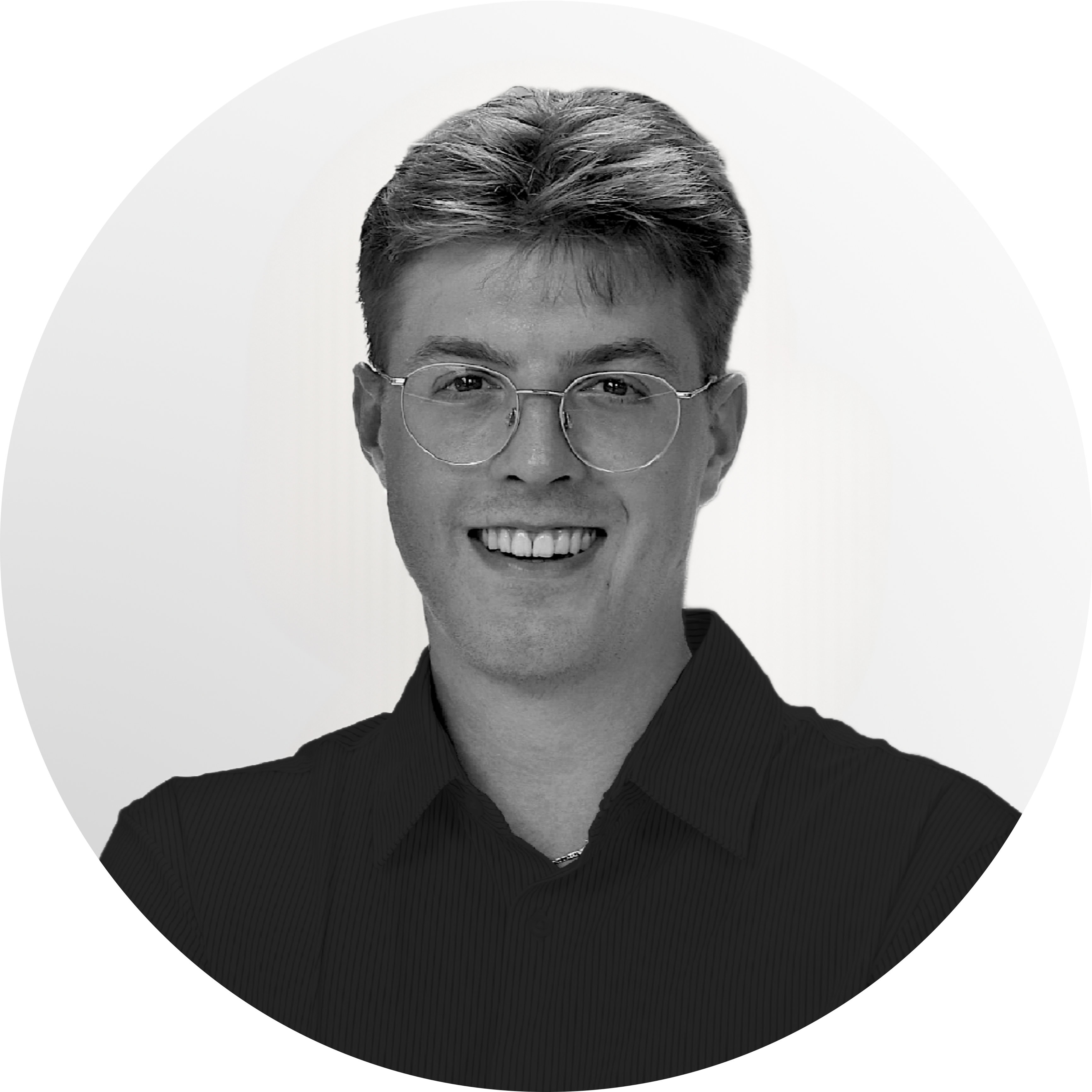 Ein Mann mit Brille, kurzen Haaren und einem Lächeln blickt direkt in die Kamera. Er trägt ein dunkelfarbiges Hemd. Der Hintergrund ist ein schlichter, hellgrauer kreisförmiger Farbverlauf.