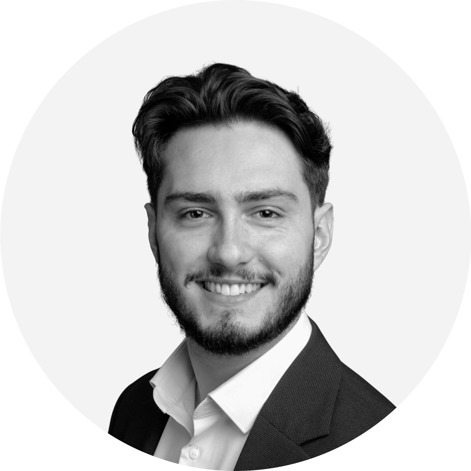 Porträt eines Mannes mit kurzen Haaren und Bart, der ein weißes Hemd und einen dunklen Blazer trägt und vor einem neutralen Hintergrund in die Kamera lächelt.