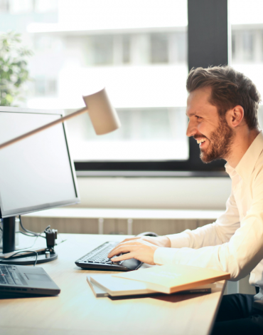 Ein Mann im weißen Hemd sitzt lächelnd an einem Schreibtisch und arbeitet in einem hellen Büro mit großen Fenstern an einem Computer.