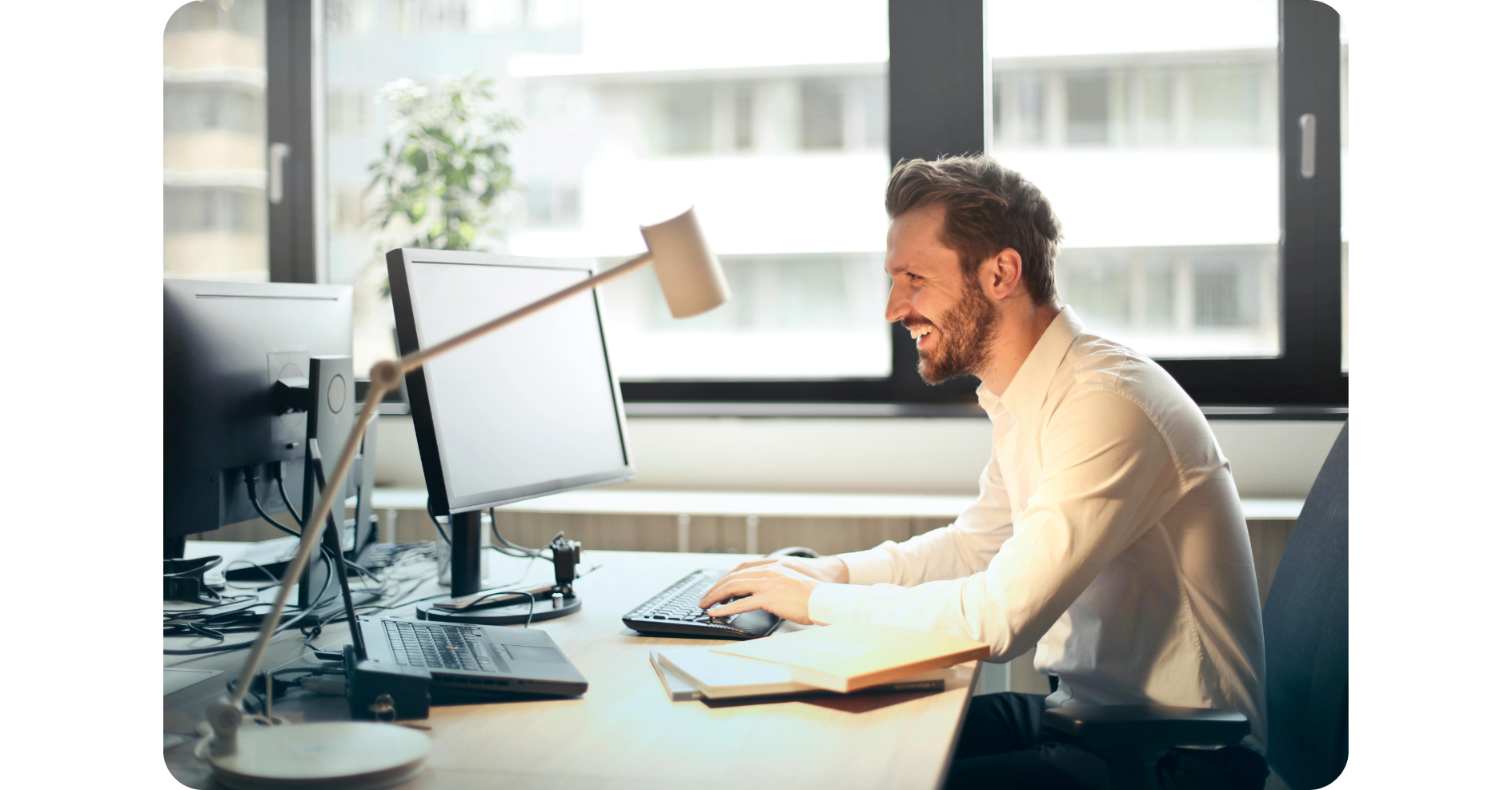 Ein Mann im weißen Hemd sitzt lächelnd an einem Schreibtisch und arbeitet in einem hellen Büro mit großen Fenstern an einem Computer.