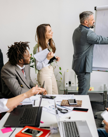 Vier Kollegen in einem modernen Büro beobachten, wie ein Mann im Anzug während einer Besprechung ein Diagramm auf ein Flipchart zeichnet; Laptops, Papiere und Telefone liegen auf dem Tisch.