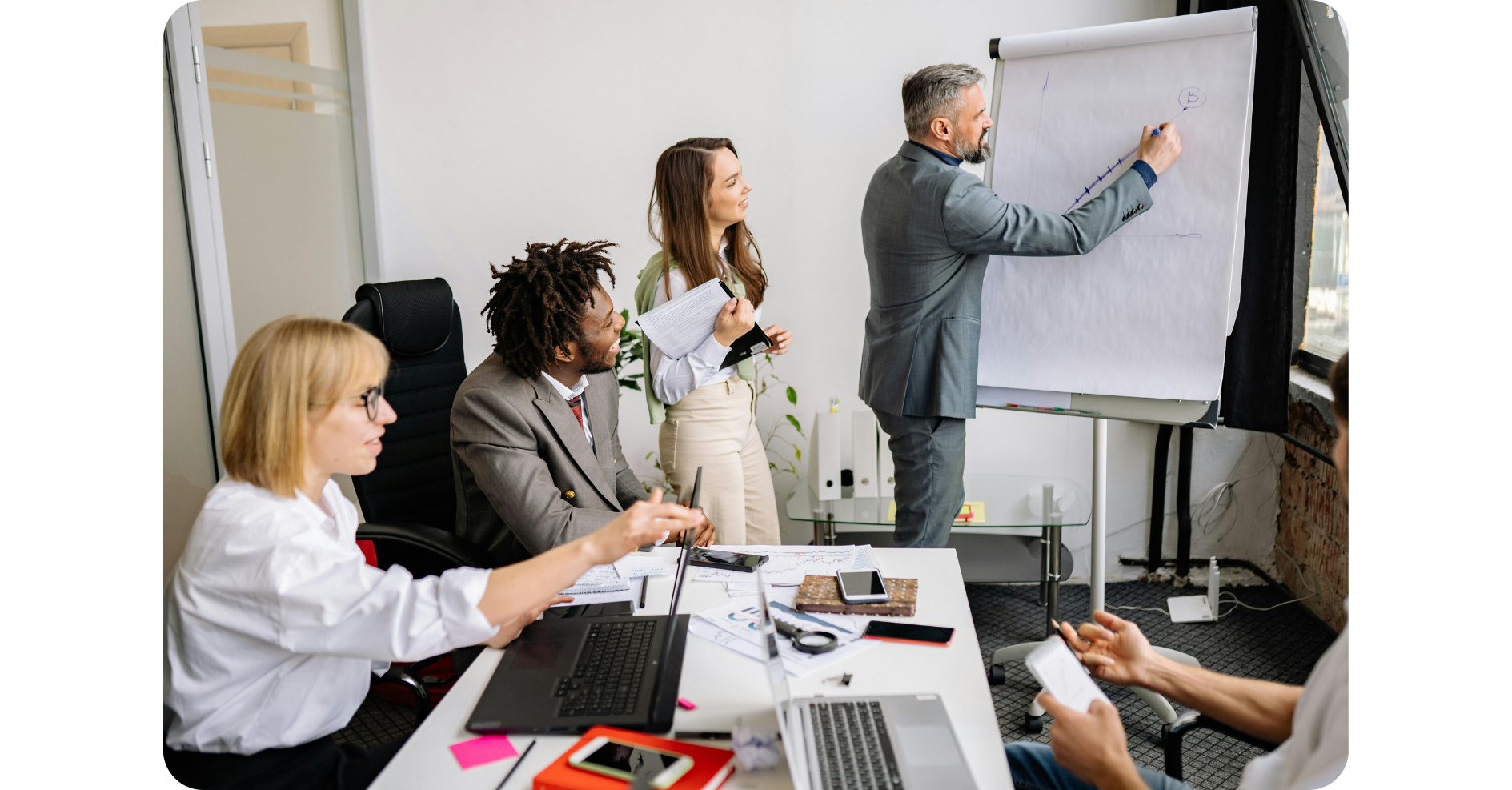 Vier Kollegen in einem modernen Büro beobachten, wie ein Mann im Anzug während einer Besprechung ein Diagramm auf ein Flipchart zeichnet; Laptops, Papiere und Telefone liegen auf dem Tisch.