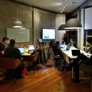Eine Gruppe von Leuten mit Laptops sitzt um einen Konferenztisch in einem modernen Büro und hört einer Frau zu, die an einem Bildschirm präsentiert.