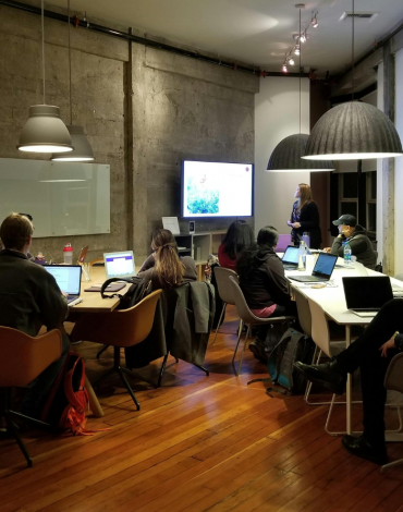 Eine Gruppe von Leuten mit Laptops sitzt um einen Konferenztisch in einem modernen Büro und hört einer Frau zu, die an einem Bildschirm präsentiert.