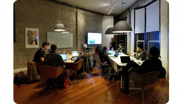 Eine Gruppe von Leuten mit Laptops sitzt um einen Konferenztisch in einem modernen Büro und hört einer Frau zu, die an einem Bildschirm präsentiert.