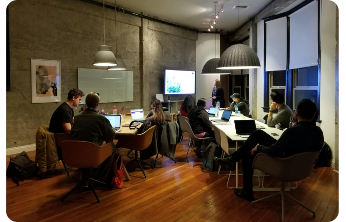 Eine Gruppe von Leuten mit Laptops sitzt um einen Konferenztisch in einem modernen Büro und hört einer Frau zu, die an einem Bildschirm präsentiert.