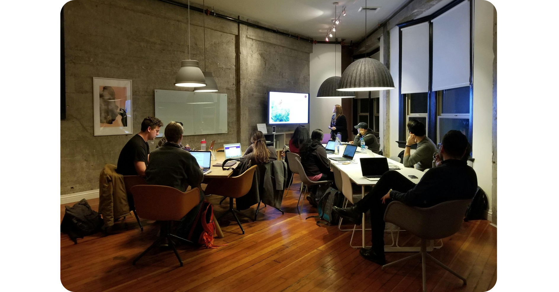 Eine Gruppe von Leuten mit Laptops sitzt um einen Konferenztisch in einem modernen Büro und hört einer Frau zu, die an einem Bildschirm präsentiert.