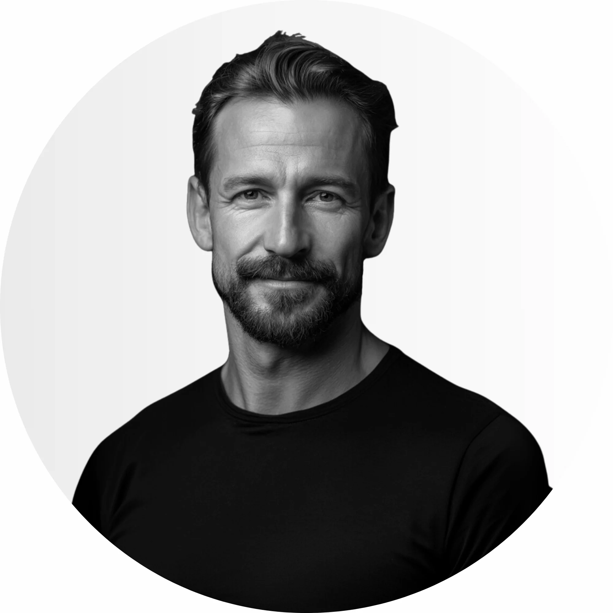 Ein Mann mit kurzen Haaren und Bart, der ein schlichtes dunkles T-Shirt tr&auml;gt, blickt nach vorne und l&auml;chelt leicht vor einem hellen, schlichten Hintergrund.