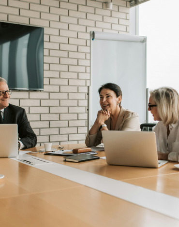 Vier Personen sitzen an einem Konferenztisch mit Laptops, Dokumenten und Kaffeetassen und führen eine Besprechung in einem modernen Büro mit großen Fenstern und einem an der Wand befestigten Fernsehbildschirm durch.