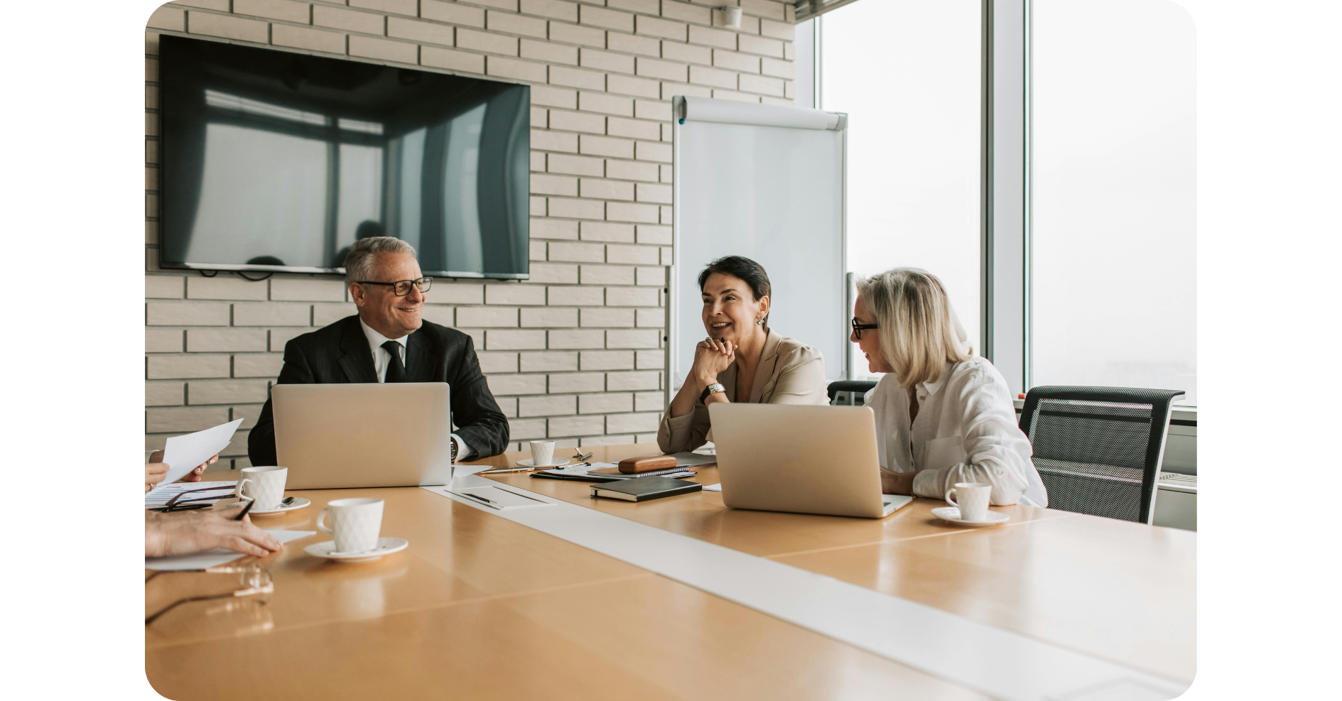 Vier Personen sitzen an einem Konferenztisch mit Laptops, Dokumenten und Kaffeetassen und führen eine Besprechung in einem modernen Büro mit großen Fenstern und einem an der Wand befestigten Fernsehbildschirm durch.