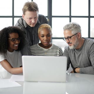 Vier Personen, zwei Männer und zwei Frauen, sitzen und stehen um einen Tisch herum und schauen gemeinsam auf einen Laptop-Bildschirm in einem hellen Büro.