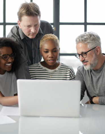 Vier Personen, zwei Männer und zwei Frauen, sitzen und stehen um einen Tisch herum und schauen gemeinsam auf einen Laptop-Bildschirm in einem hellen Büro.