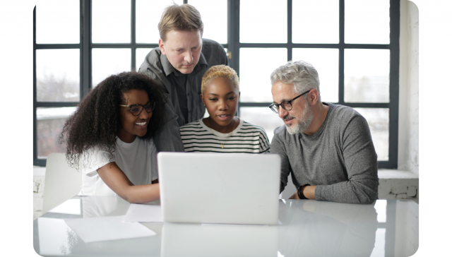 Vier Personen, zwei Männer und zwei Frauen, sitzen und stehen um einen Tisch herum und schauen gemeinsam auf einen Laptop-Bildschirm in einem hellen Büro.