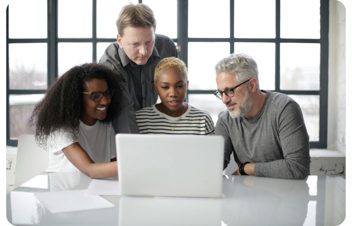 Vier Personen, zwei Männer und zwei Frauen, sitzen und stehen um einen Tisch herum und schauen gemeinsam auf einen Laptop-Bildschirm in einem hellen Büro.