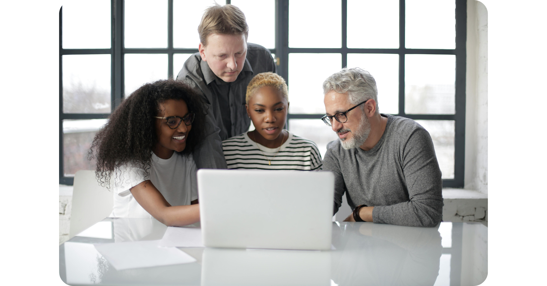 Vier Personen, zwei Männer und zwei Frauen, sitzen und stehen um einen Tisch herum und schauen gemeinsam auf einen Laptop-Bildschirm in einem hellen Büro.