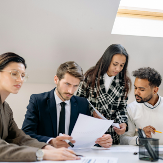 Vier Fachleute sitzen und stehen um einen Tisch herum, prüfen Dokumente und besprechen die Arbeit in einem hellen Büro mit Tageslicht durch ein Oberlicht.