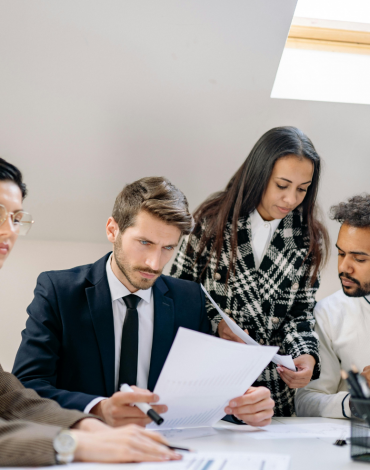 Vier Fachleute sitzen und stehen um einen Tisch herum, prüfen Dokumente und besprechen die Arbeit in einem hellen Büro mit Tageslicht durch ein Oberlicht.