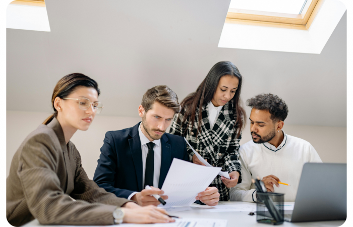 Vier Fachleute sitzen und stehen um einen Tisch herum, prüfen Dokumente und besprechen die Arbeit in einem hellen Büro mit Tageslicht durch ein Oberlicht.