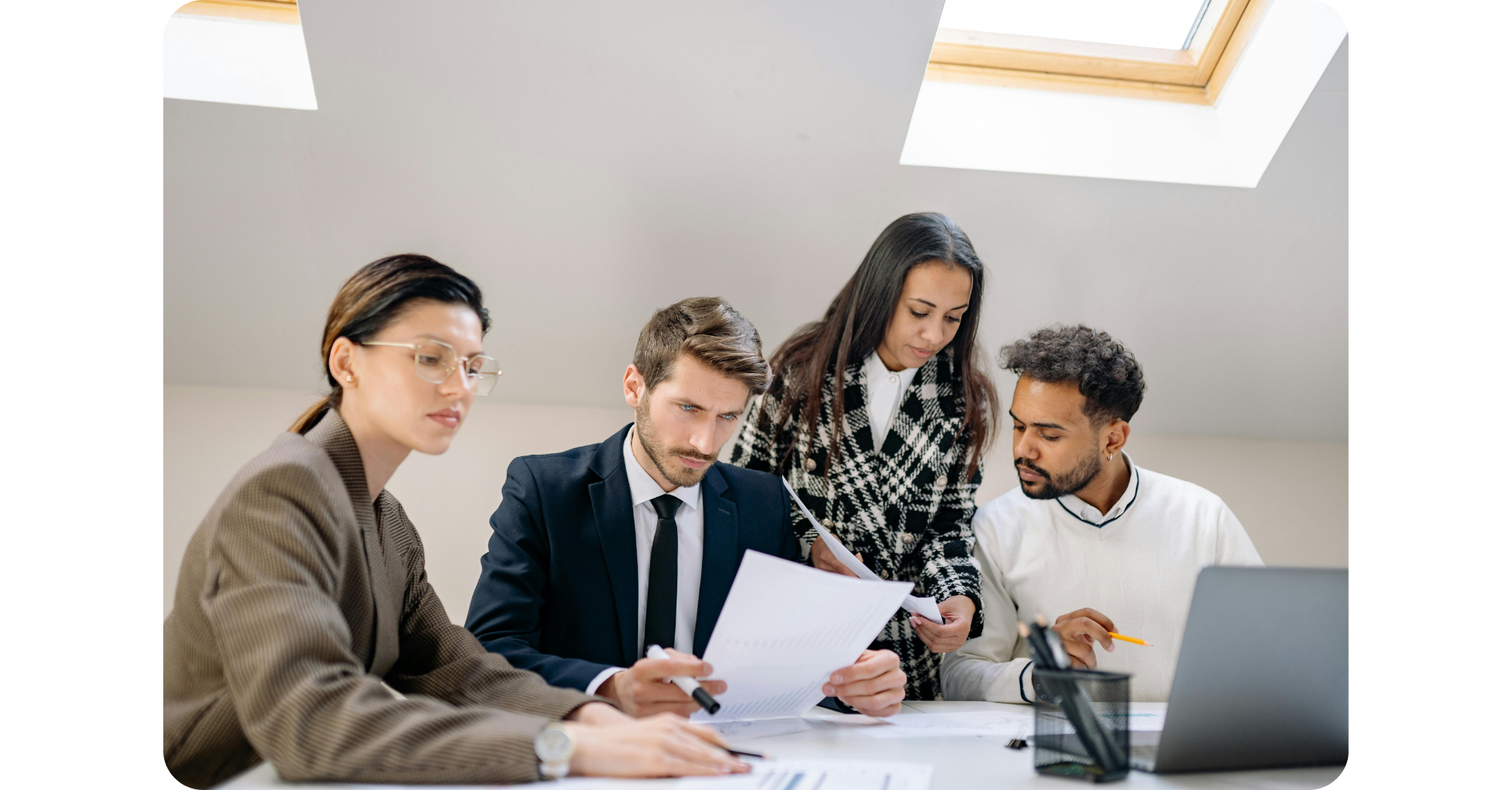 Vier Fachleute sitzen und stehen um einen Tisch herum, prüfen Dokumente und besprechen die Arbeit in einem hellen Büro mit Tageslicht durch ein Oberlicht.
