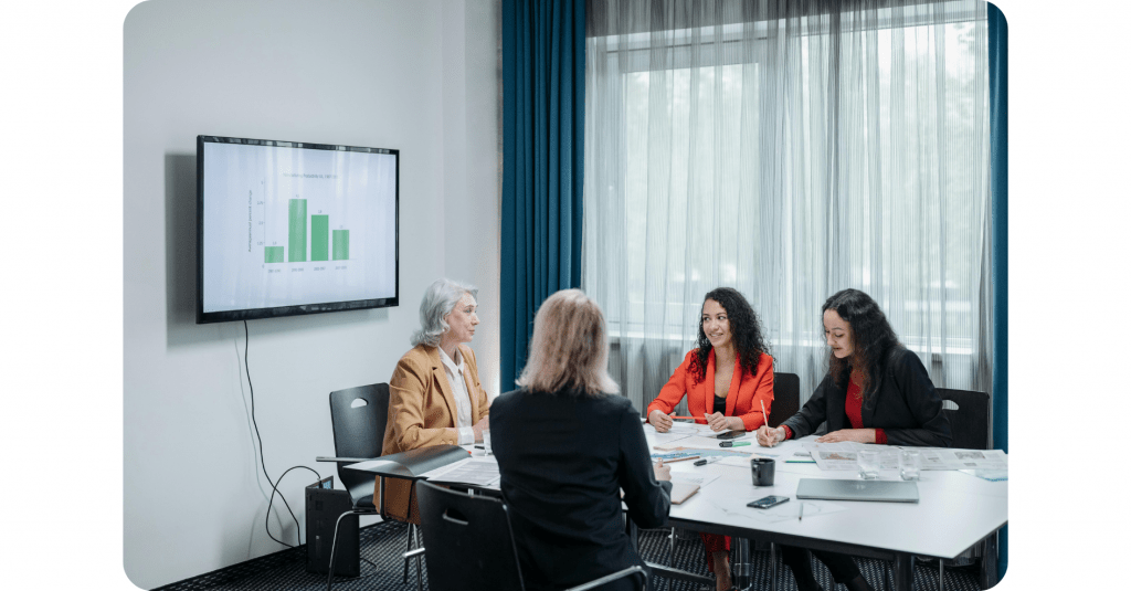 Vier Frauen sitzen in einem Besprechungsraum mit Unterlagen und einem Laptop um einen Konferenztisch, während auf einem Wandbildschirm ein Balkendiagramm angezeigt wird.