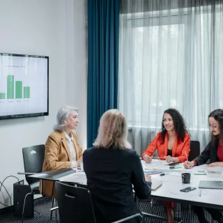 Four women sit around a conference table in a meeting room with documents and a laptop, while a bar chart is displayed on a wall screen.