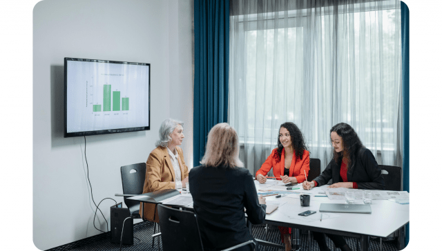 Vier Frauen sitzen in einem Besprechungsraum mit Unterlagen und einem Laptop um einen Konferenztisch, während auf einem Wandbildschirm ein Balkendiagramm angezeigt wird.