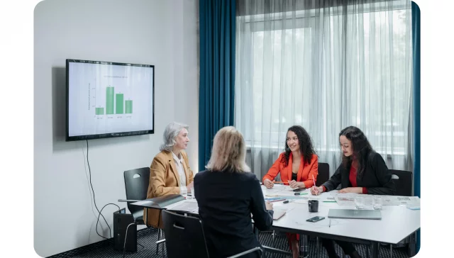 Four women sit around a conference table in a meeting room with documents and a laptop, while a bar chart is displayed on a wall screen.