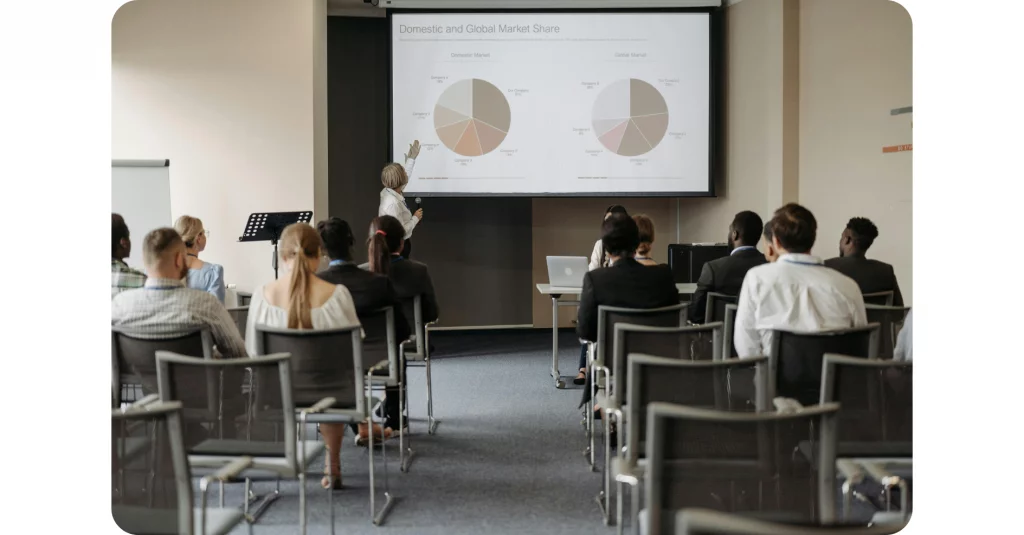 A speaker points to pie charts on a screen labeled "Domestic and Global Market Share" while the audience sits in a conference room and listens.