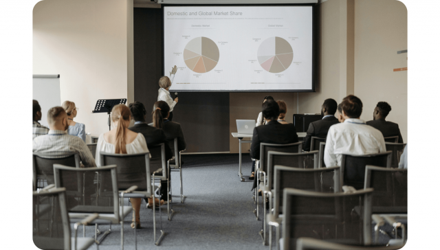 Ein Redner zeigt auf Kuchendiagramme auf einem Bildschirm mit der Aufschrift "Inländischer und globaler Marktanteil", während das Publikum in einem Konferenzraum sitzt und zuhört.