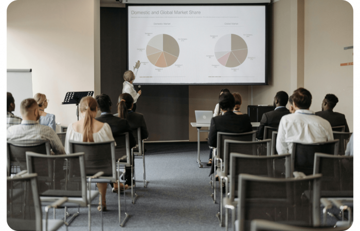 Ein Redner zeigt auf Kuchendiagramme auf einem Bildschirm mit der Aufschrift "Inländischer und globaler Marktanteil", während das Publikum in einem Konferenzraum sitzt und zuhört.